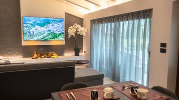 A modern living room with a flat-screen TV, a fireplace, a dining area with a table set for two, and a large window with curtains.