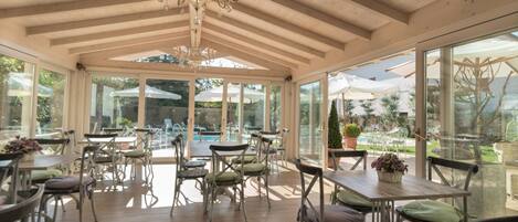 A covered outdoor dining area with wooden tables and chairs, a chandelier, and large windows overlooking a pool and garden.