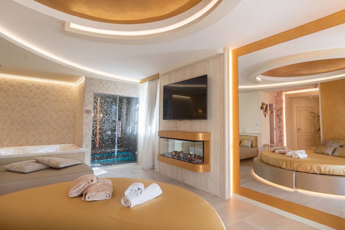 Quarto de hotel moderno com uma cama grande, televisão integrada, box de chuveiro em vidro e um espaço de estar com sofá e uma mesa pequena.