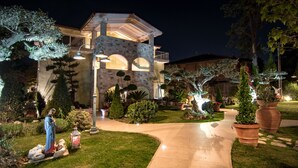 A well-lit, modern house with a stone facade, arched windows, and a landscaped garden with potted plants and a statue.