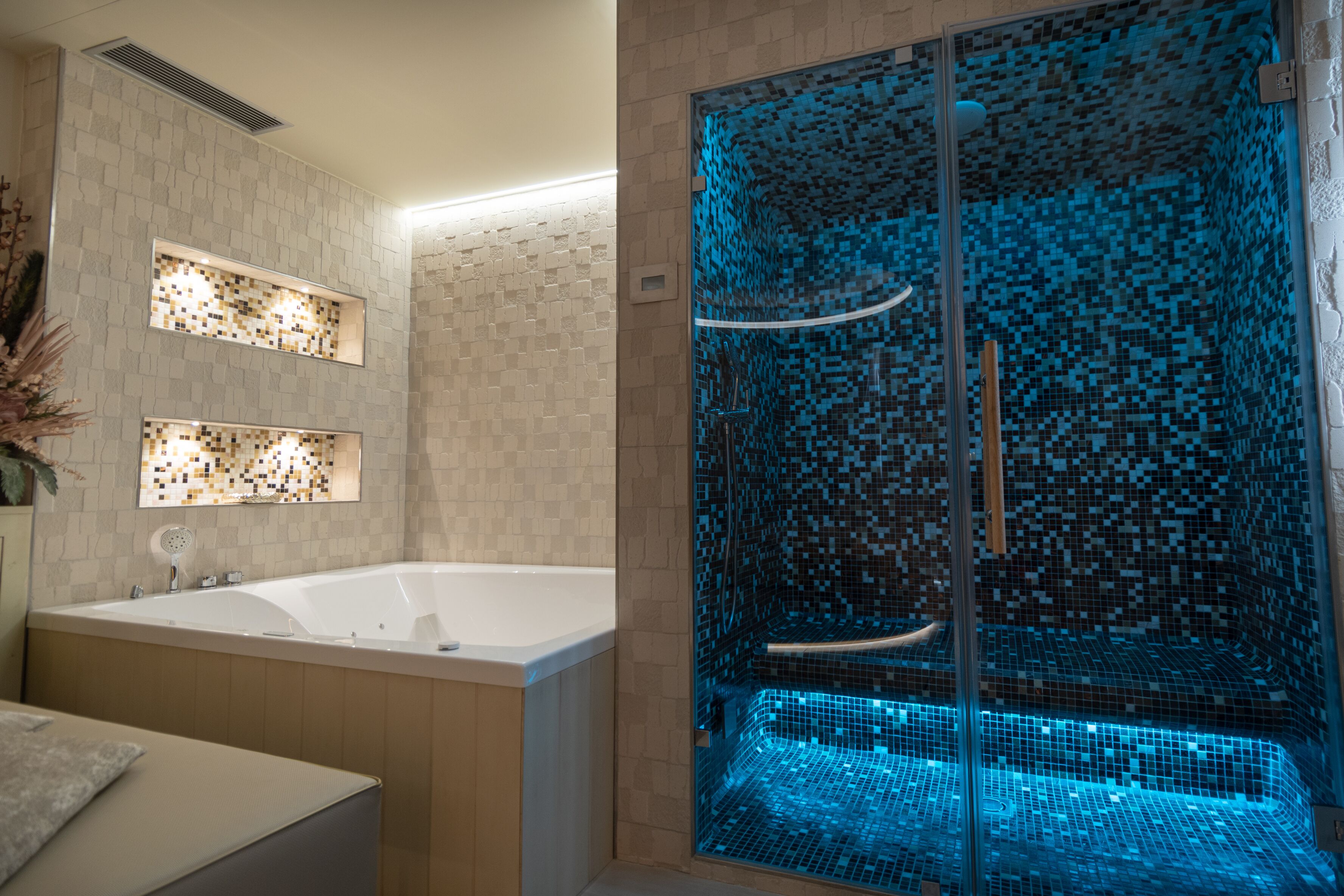 A modern bathroom with a large bathtub, a glass-enclosed shower with blue mosaic tiles, and two illuminated wall panels.