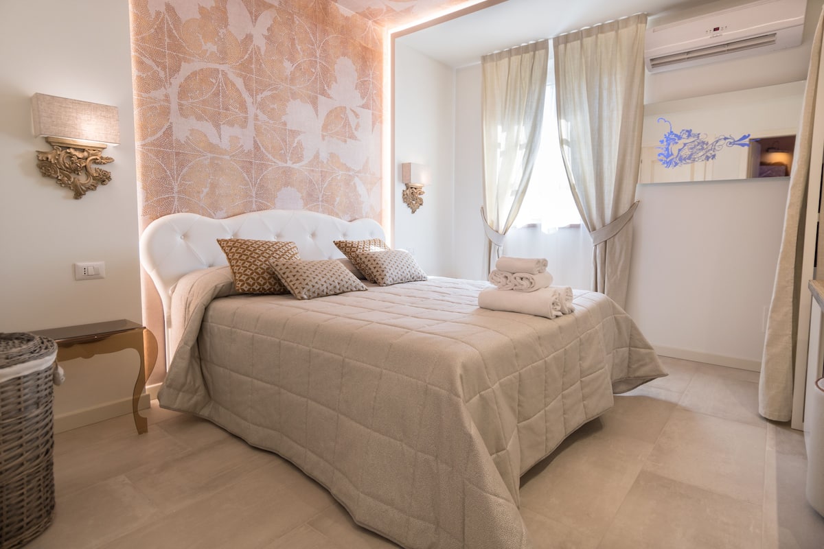 Quarto com cama grande, mesa de cabeceira, luminária na parede e painel decorativo.