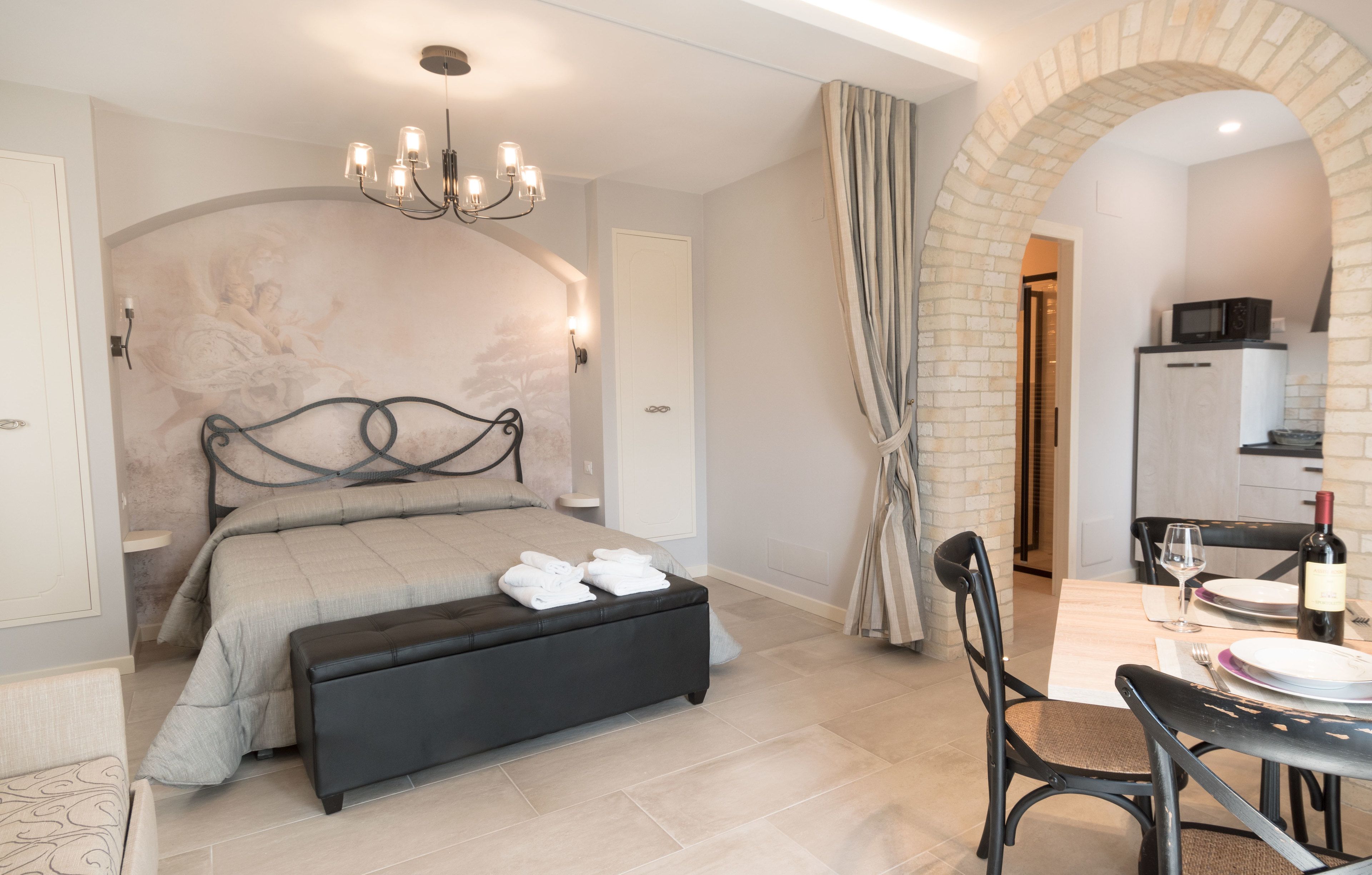 Un dormitorio con cama, un banco, una mesa de comedor con sillas y una zona de cocina visible a través de un arco.
