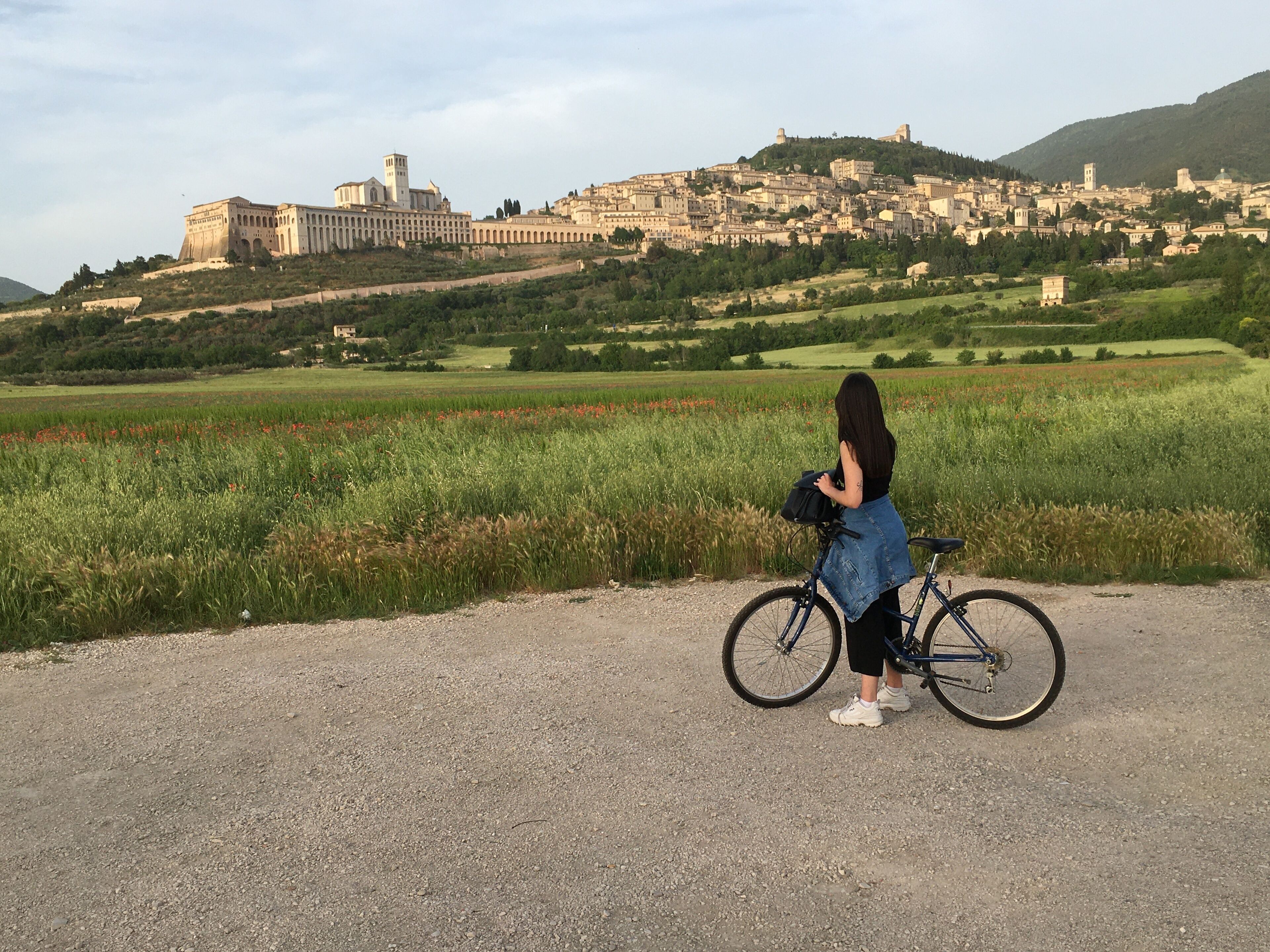 A person standing next to a bicycle on a gravel path with a view of a historic cityscape in the background.