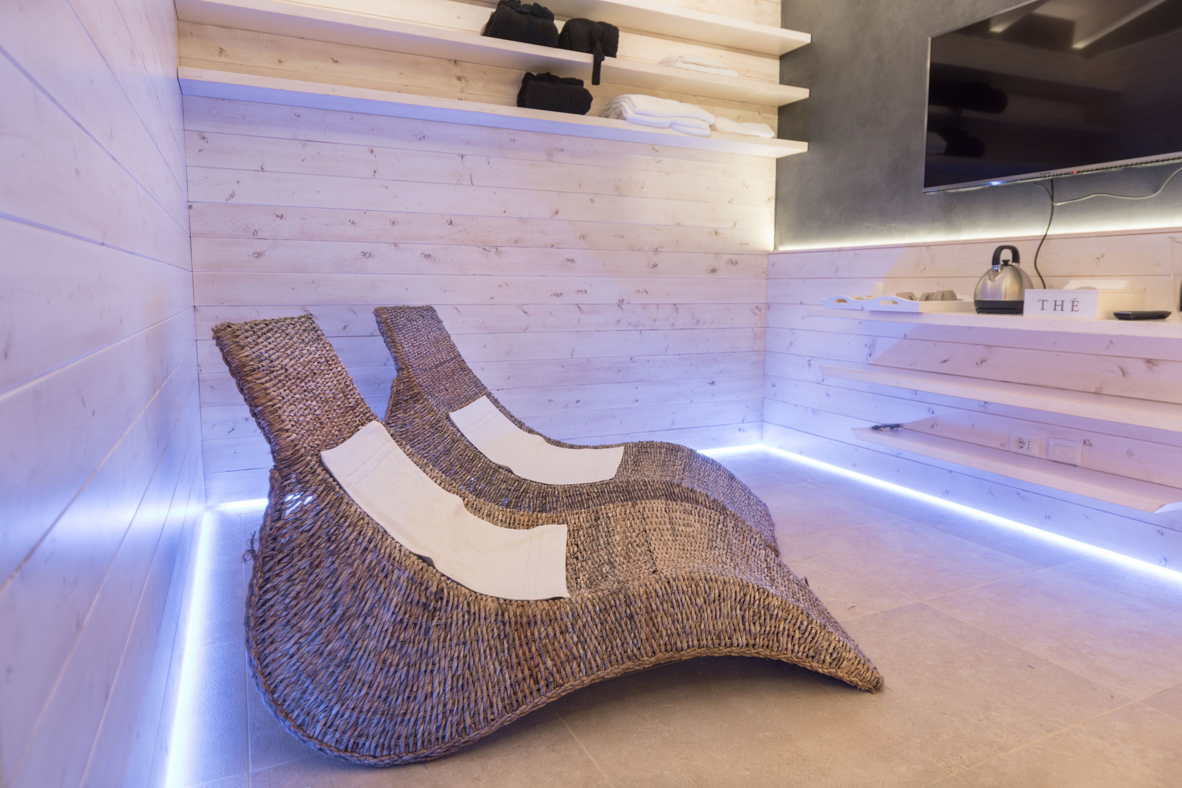 A modern room with a woven chair, wooden shelves, a flat-screen TV, and a kettle on a shelf.