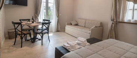 A hotel room with a bed, a sofa, a dining table with chairs, a TV mounted on the wall, and an air conditioning unit.