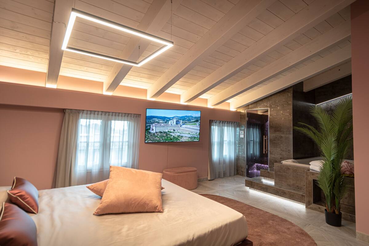 Quarto de hotel moderno com uma cama grande, televisão de tela plana, área de estar com lareira e vista para a paisagem urbana.