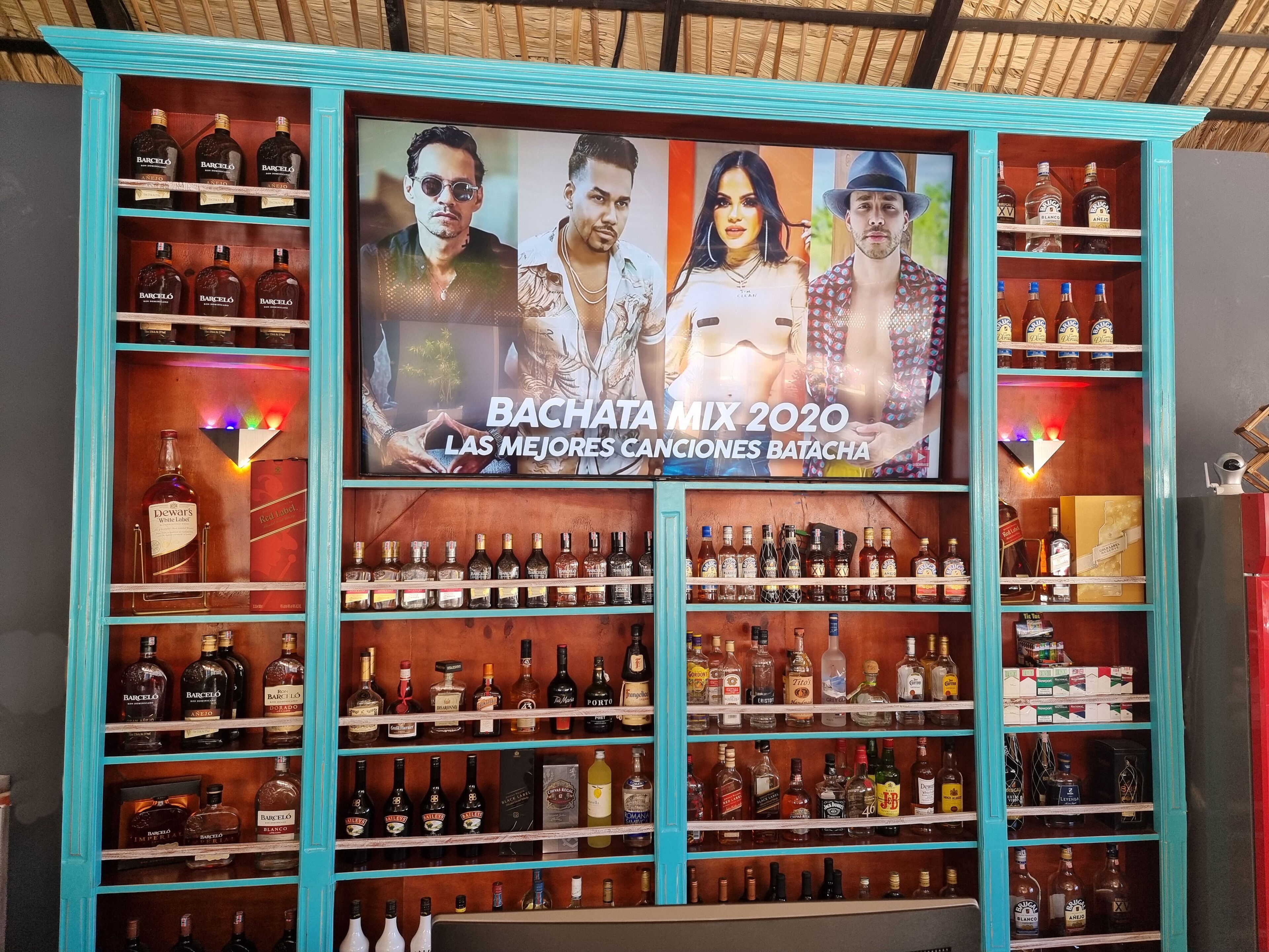 Een bar met een groot scherm waarop 'Bachata Mix 2020' draait en kasten vol met verschillende drankflessen.