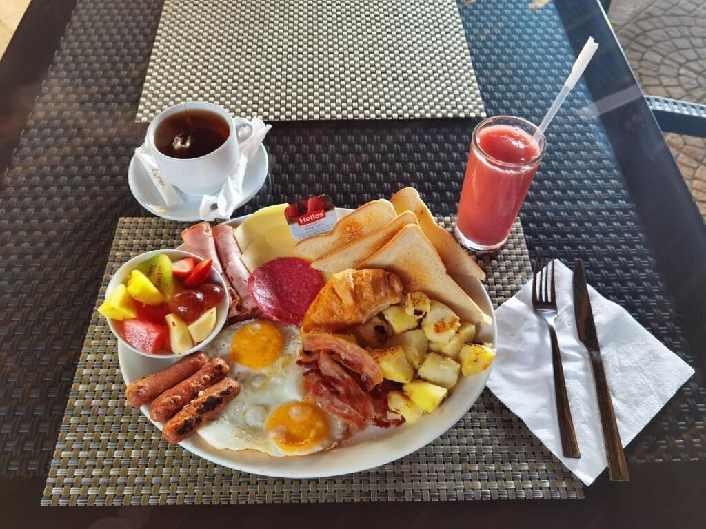 Un plato de desayuno con huevos, tocino, salchichas, tostadas y fruta, junto con una taza de café y un vaso de jugo sobre una mesa.