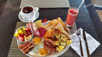 Un plato de desayuno con huevos, tocino, salchichas, tostadas y fruta, junto con una taza de café y un vaso de jugo sobre una mesa.