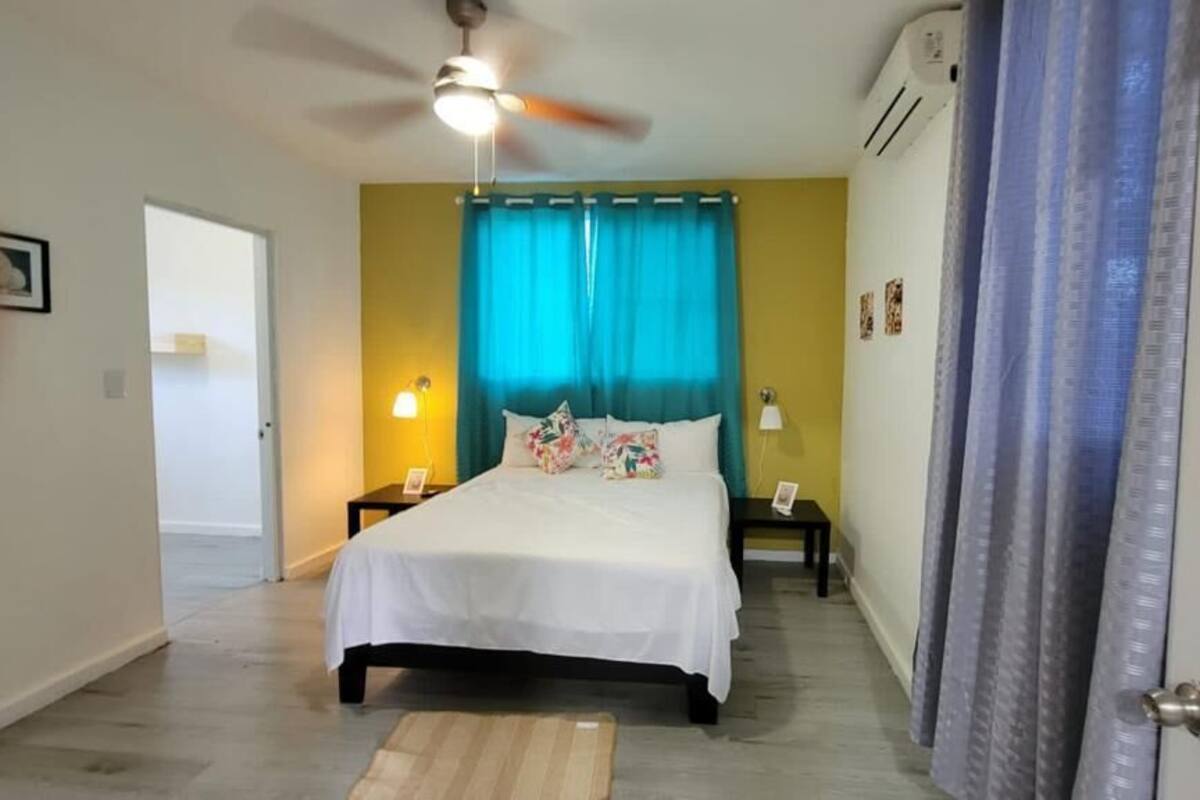 Ein Schlafzimmer mit einem Bett, einem Deckenventilator, einem Nachttisch, einer Lampe und einer Tür.