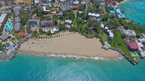 Luchtbeeld van een kustresort met een zandstrand, helder turquoise water en verschillende gebouwen, waaronder hotels en villa's.