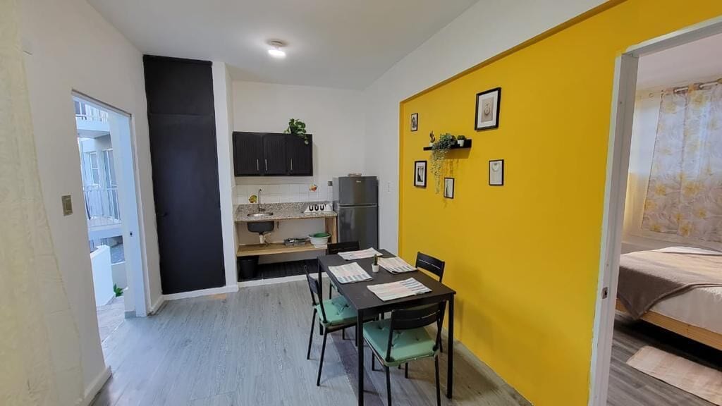 A compact kitchen with dark cabinets, a dining table with chairs, and a yellow accent wall with framed pictures.