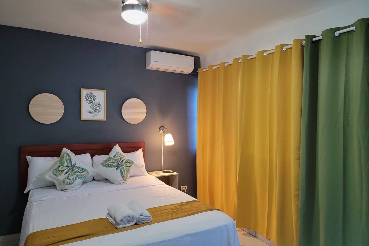 Ein Hotelzimmer mit einem Bett, einem Nachttisch, einer Klimaanlage und einem Fenster mit gelben Vorhängen.