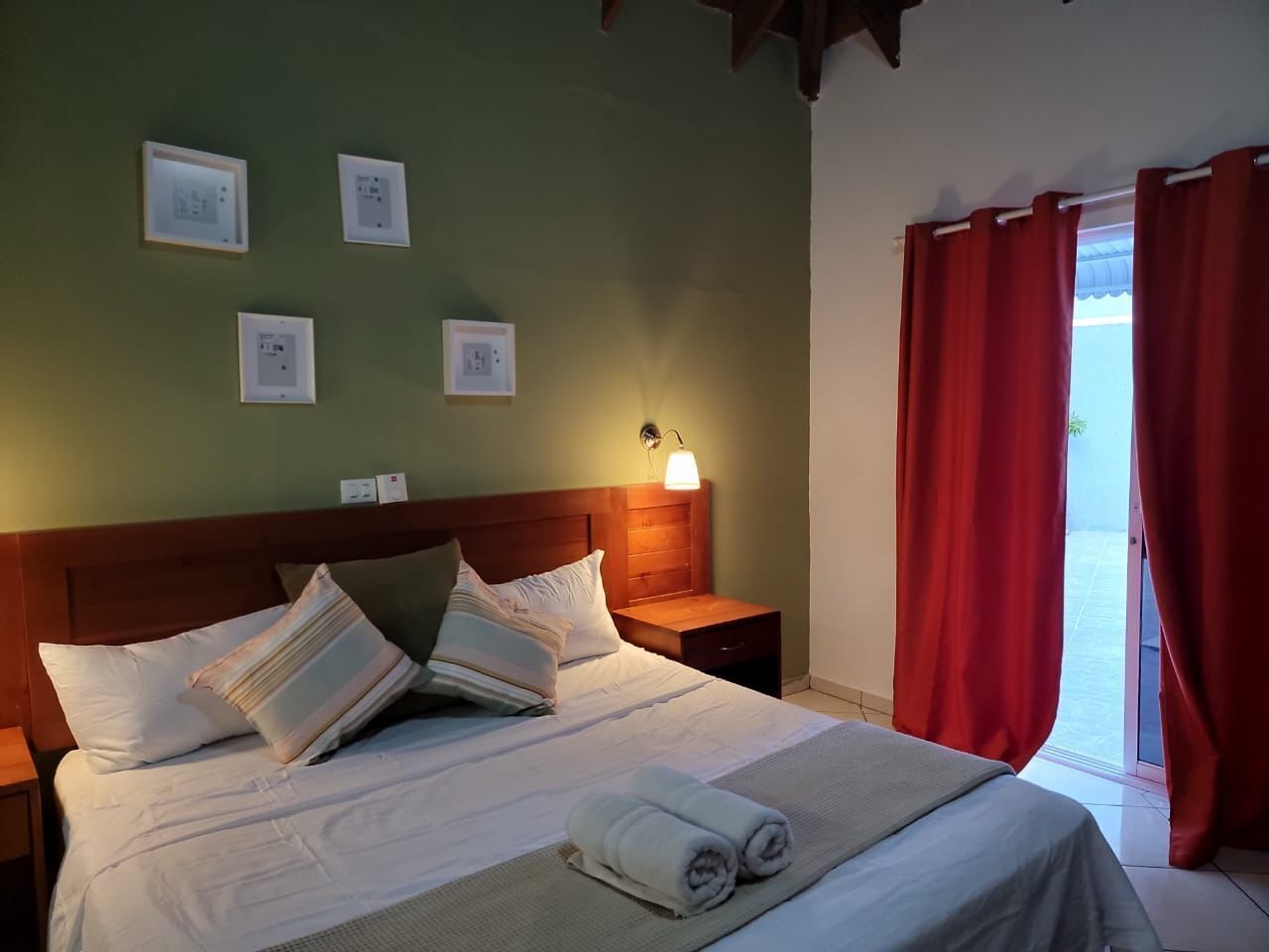A neatly made bed with pillows, a bedside lamp, and a nightstand. There are framed pictures on the wall and a window with red curtains.