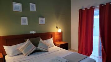 A neatly made bed with pillows, a bedside lamp, and a nightstand. There are framed pictures on the wall and a window with red curtains.