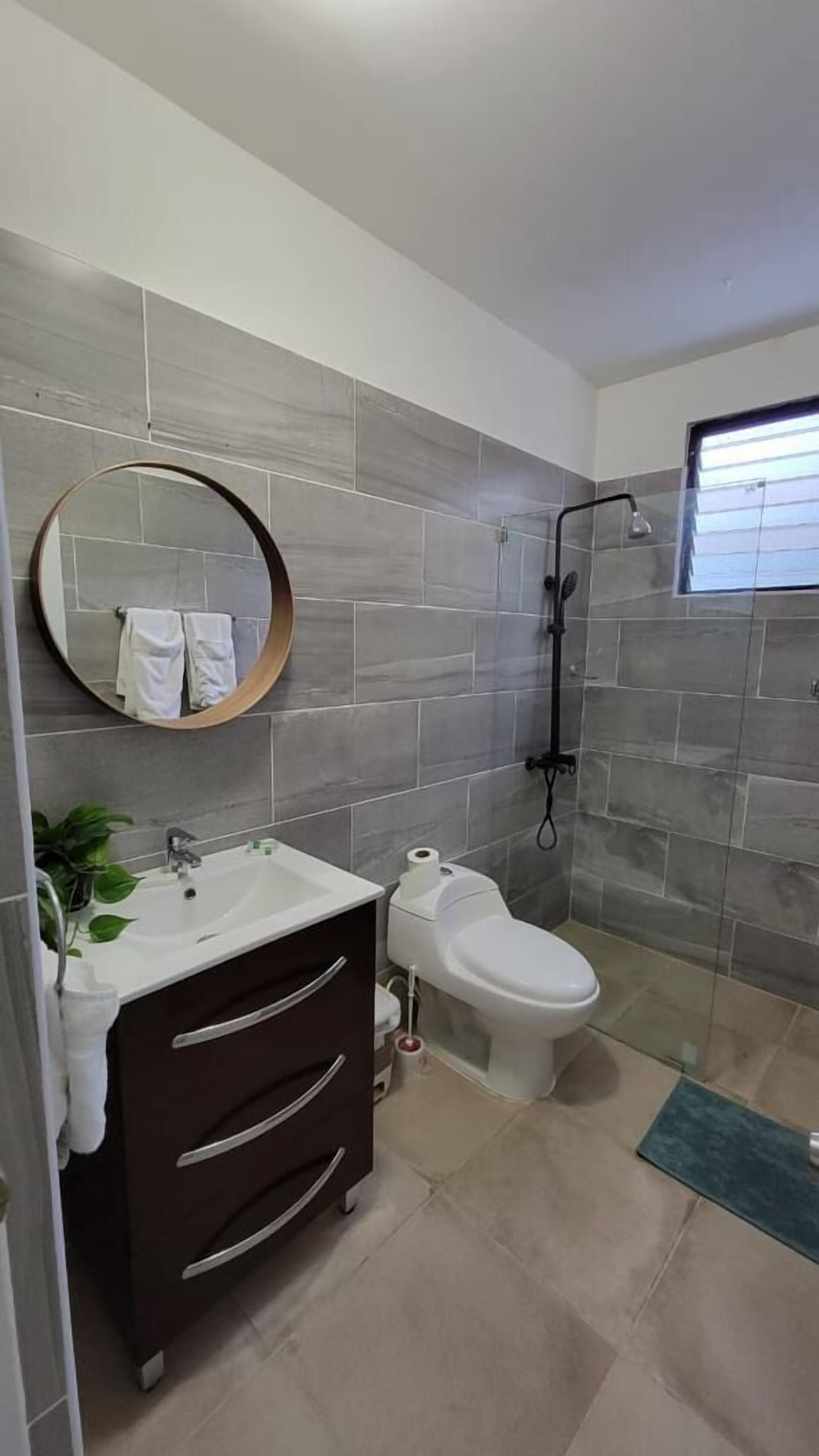 A modern bathroom with grey tiles, a round mirror, a white sink, a toilet, and a shower area.