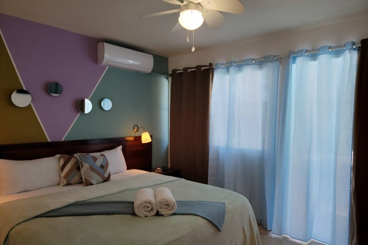 Ein Hotelzimmer mit Bett, Deckenventilator, Klimaanlage, Nachttisch, Lampe und einem Fenster mit Vorhängen.