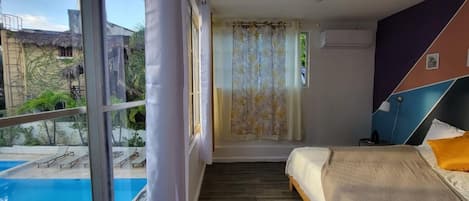 A bedroom with a bed, a window with a view of a swimming pool, and a curtain.