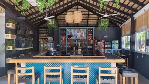 Een bar met een houten toonbank, drie houten barkrukken, plafondventilatoren en hangplanten.