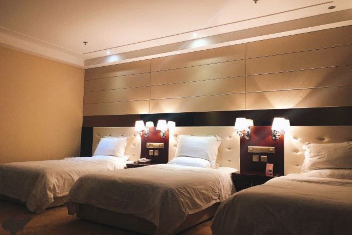 A hotel room with two beds, a large padded headboard, and wall-mounted lamps.
