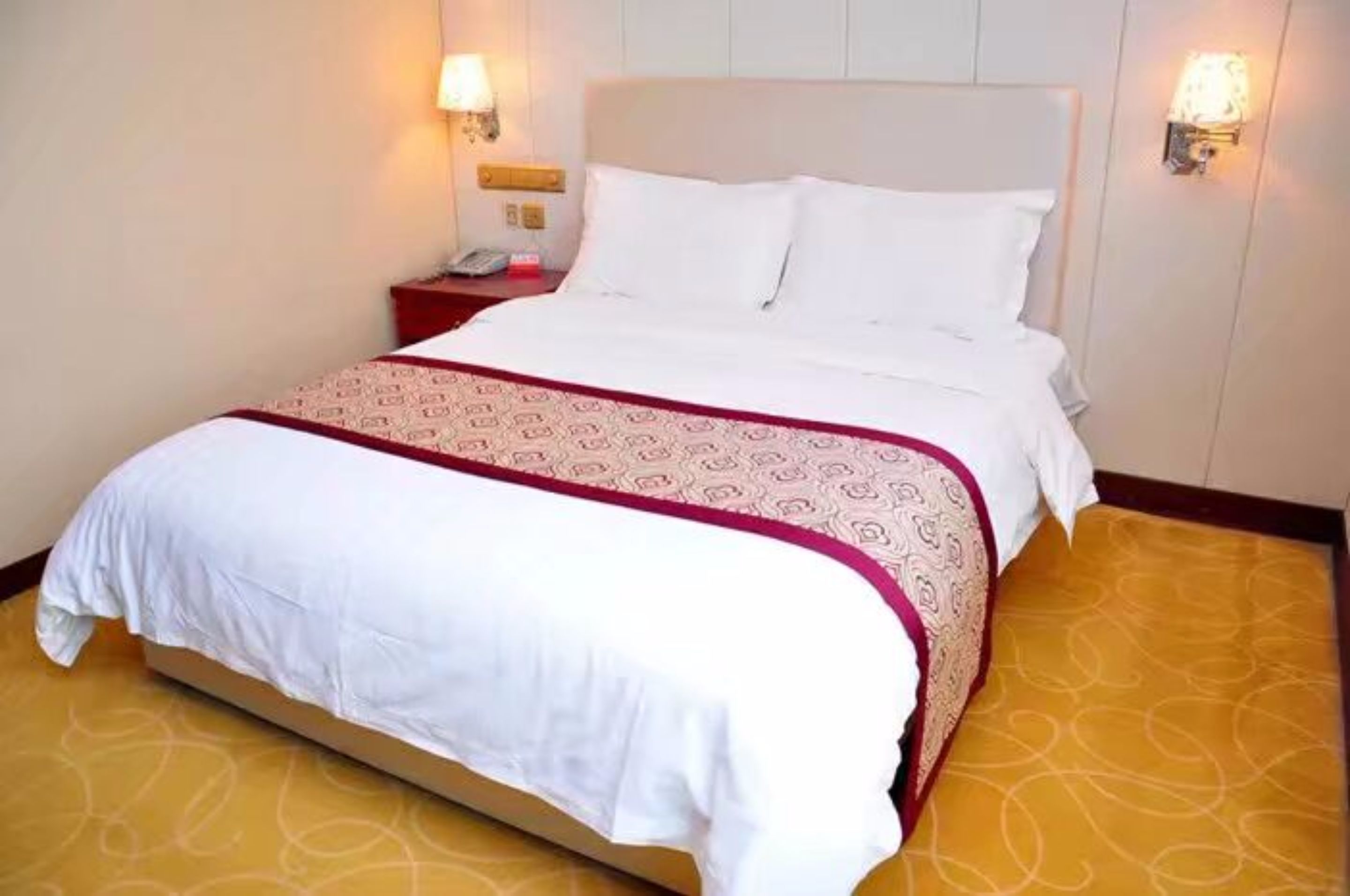 Cama bem arrumada com roupa de cama branca e colcha vermelha e branca estampada. Há duas mesas de cabeceira com abajures, e o quarto tem um tapete amarelo com padrão discreto.