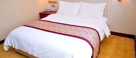 Cama bem arrumada com roupa de cama branca e colcha vermelha e branca estampada. Há duas mesas de cabeceira com abajures, e o quarto tem um tapete amarelo com padrão discreto.