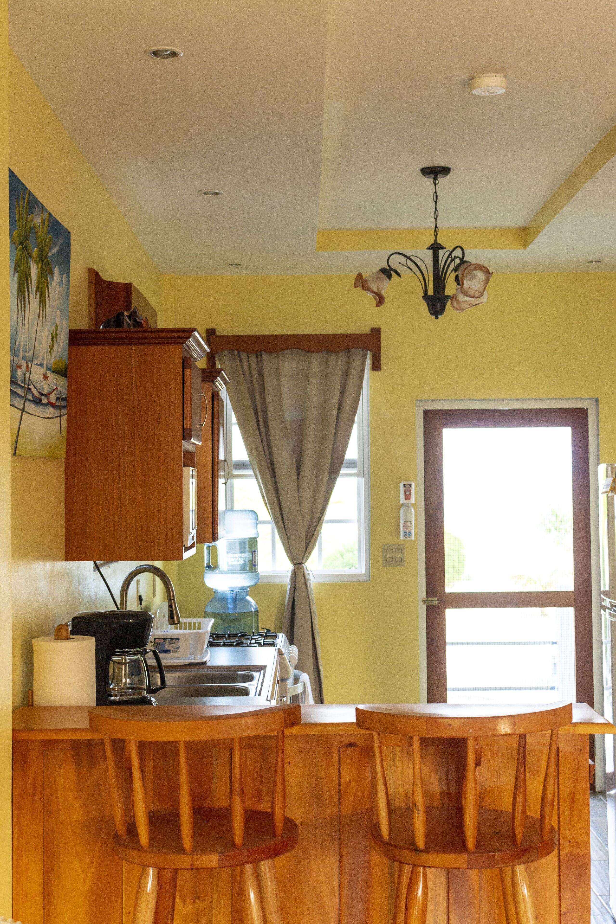 A kitchen with wooden cabinets, a bar area with stools, a water dispenser, and a window with curtains.
