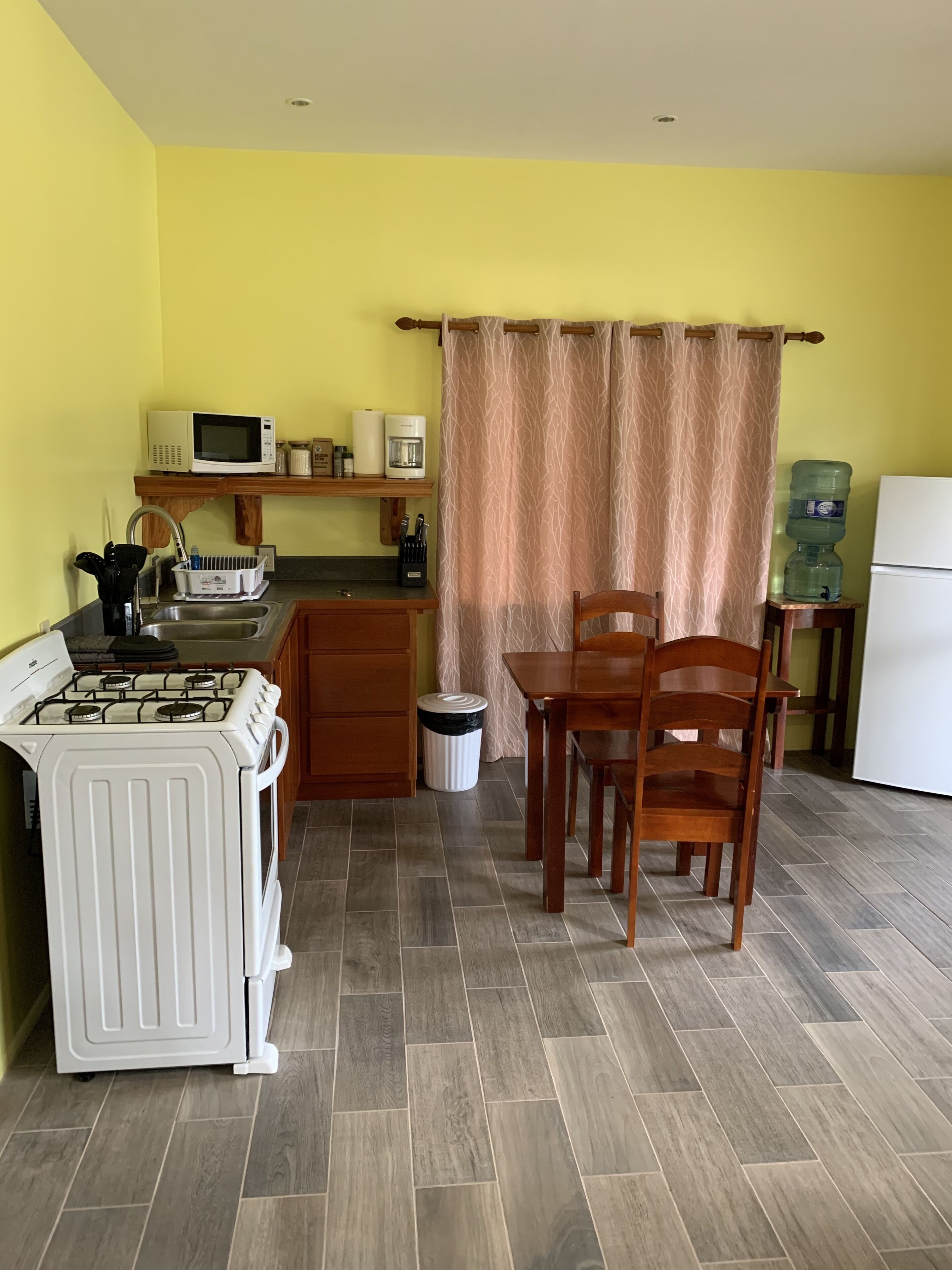 Cocina compacta con estufa a gas, microondas y cafetera. Cuenta con una mesa de comedor con sillas, dispensador de agua y un basurero.