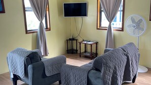 A room with two sofas covered in quilts, a flat-screen TV mounted on the wall, a standing fan, and a small table with a remote control.
