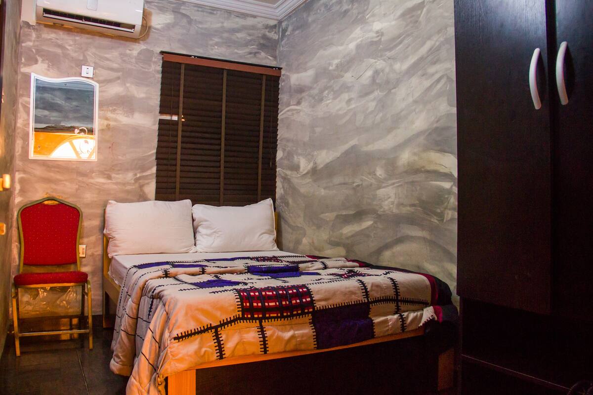 A bedroom with a bed, a chair, a wardrobe, and a wall-mounted air conditioner.