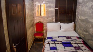 A small room with a bed, a chair, a window with blinds, and an air conditioning unit on the wall. - Glass House Suites Annex (Lagos)
