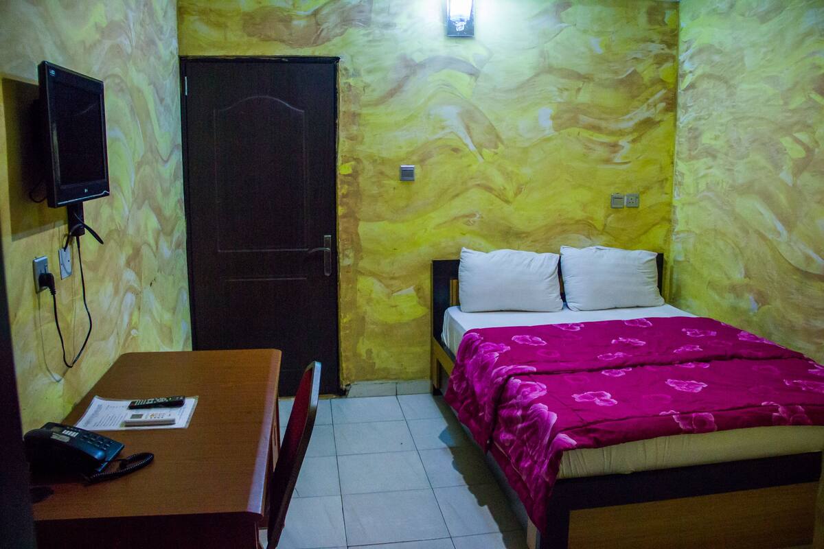 A room with a bed, a desk, a chair, a TV mounted on the wall, and a door.