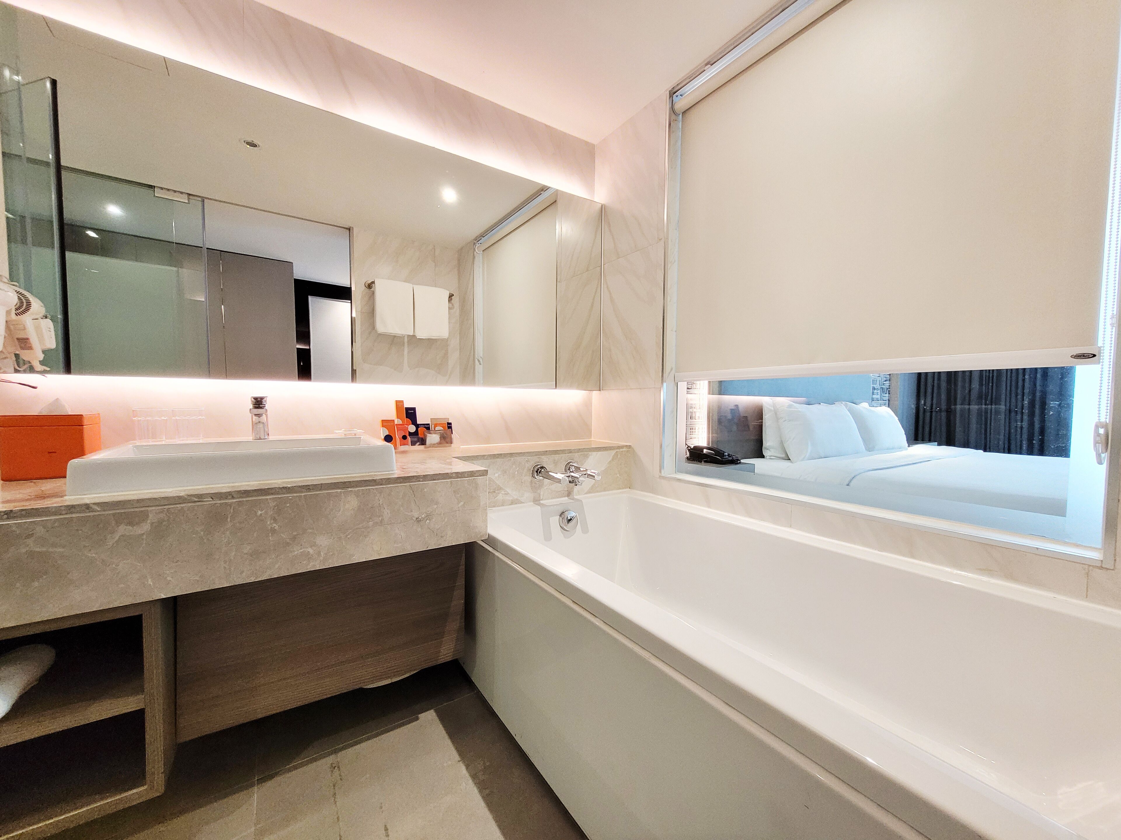 Een moderne badkamer met een grote spiegel, een badkuip en een raam met uitzicht op een bed.