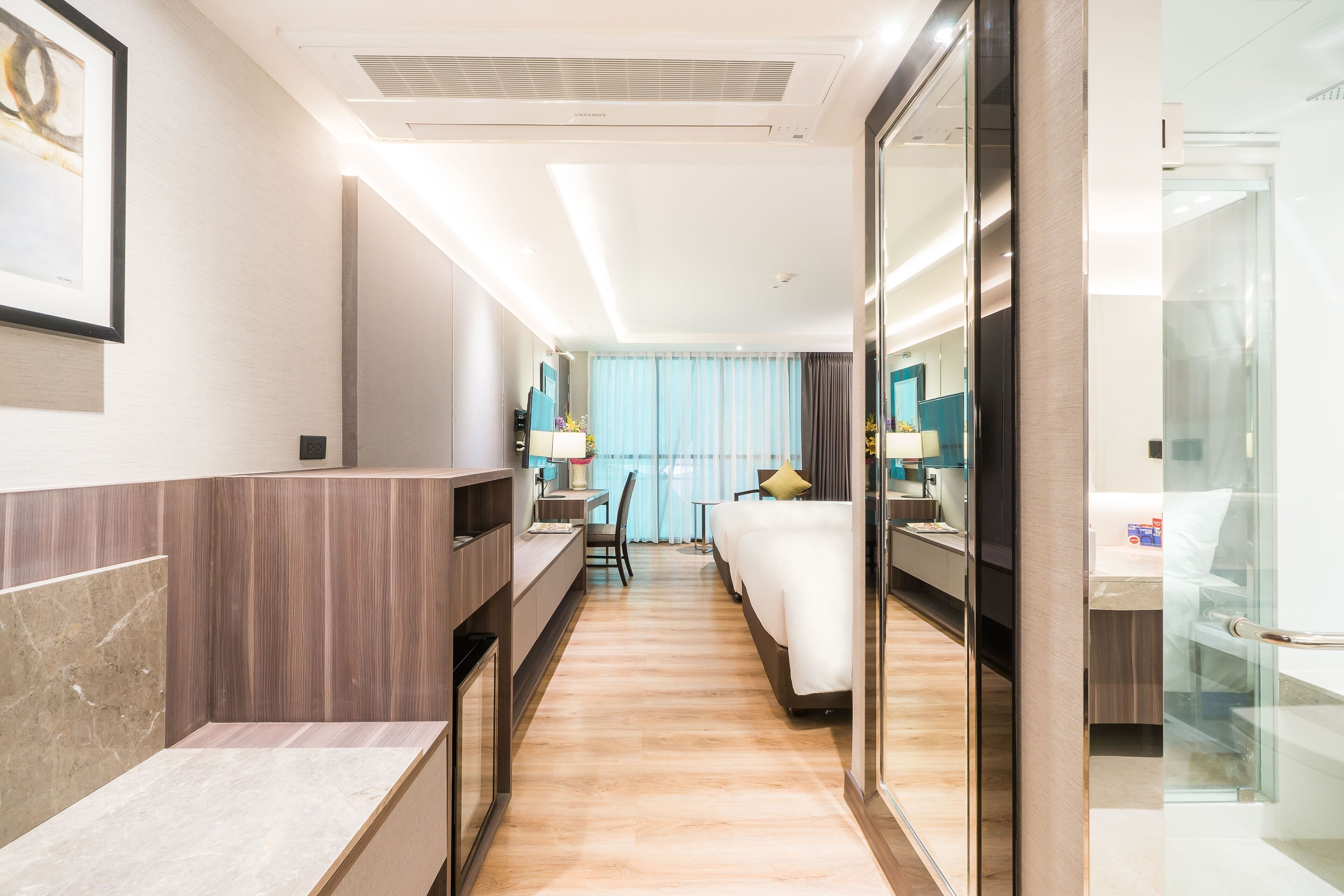 Una habitación de hotel moderna con cama, mesita de noche y escritorio con silla. Hay un espejo grande en una pared y una ventana con cortinas.