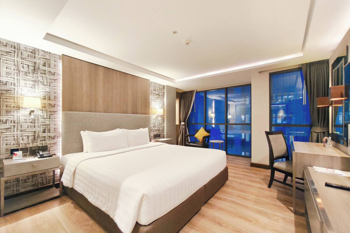 A modern hotel room with a large bed, a desk, and a view of the city through large windows.
