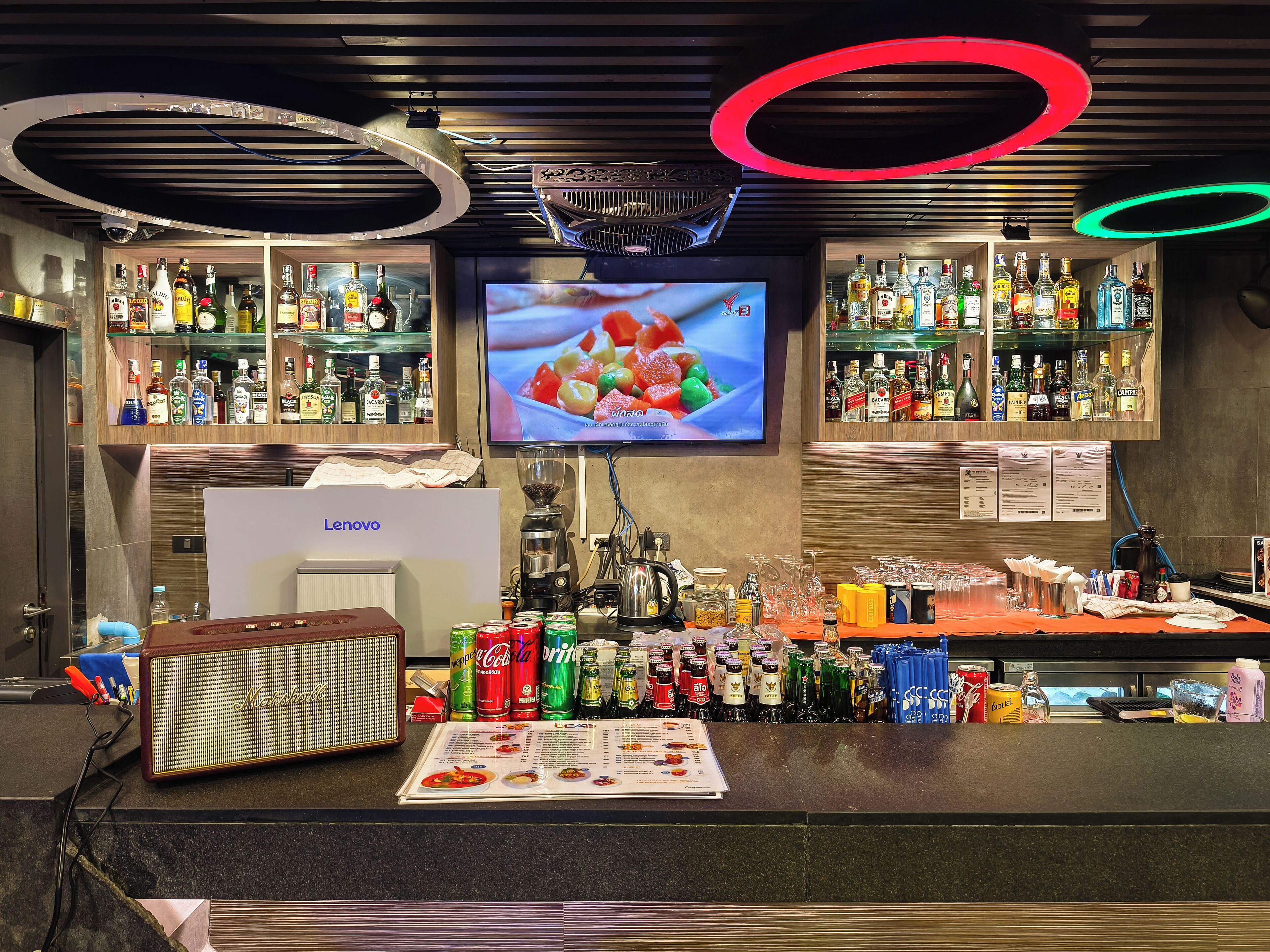 Una zona de bar con estanterías llenas de botellas de licores, una pantalla de televisión, una cafetera y un menú.