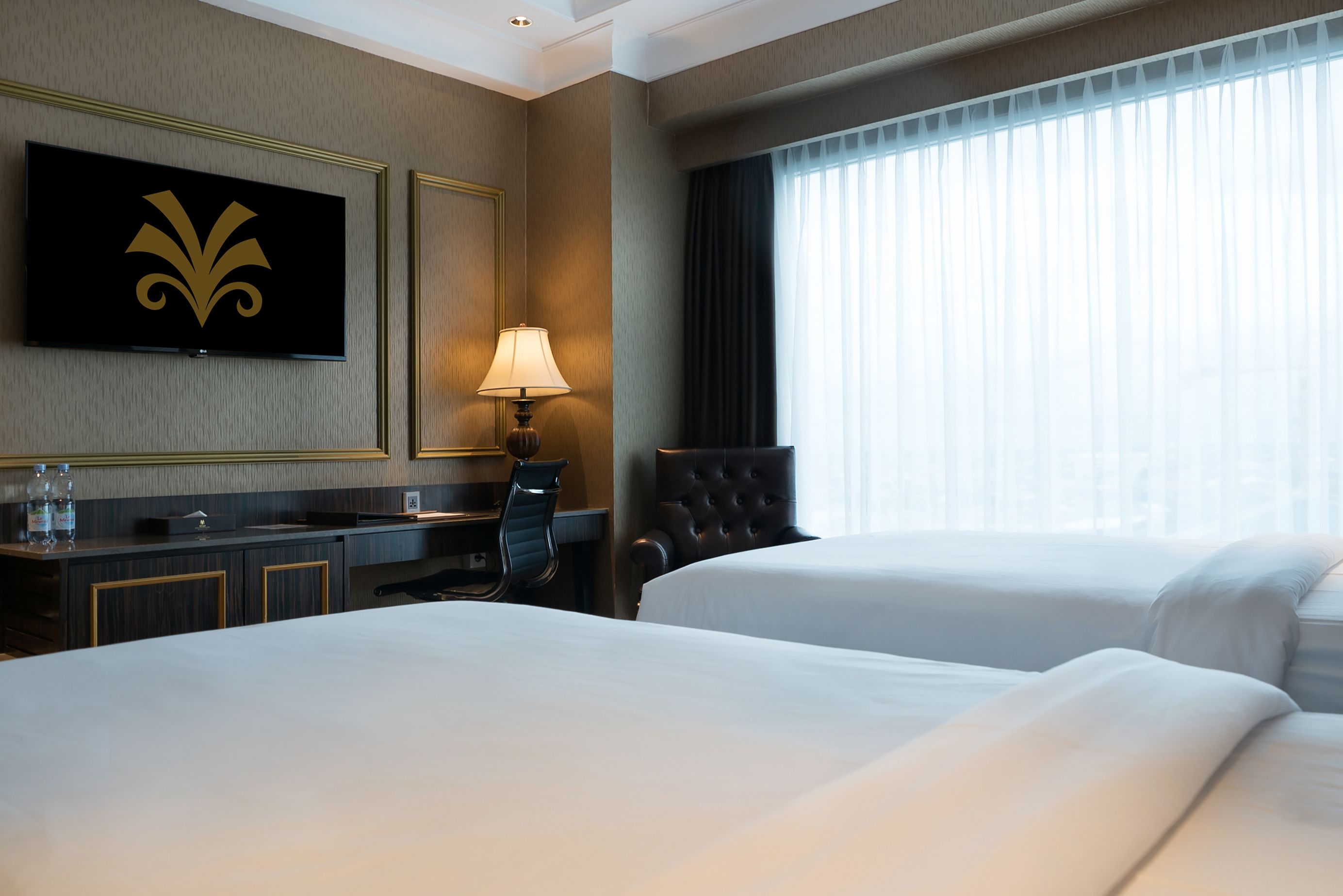 A hotel room with two beds, a desk with a lamp, a television, and a large window with curtains.