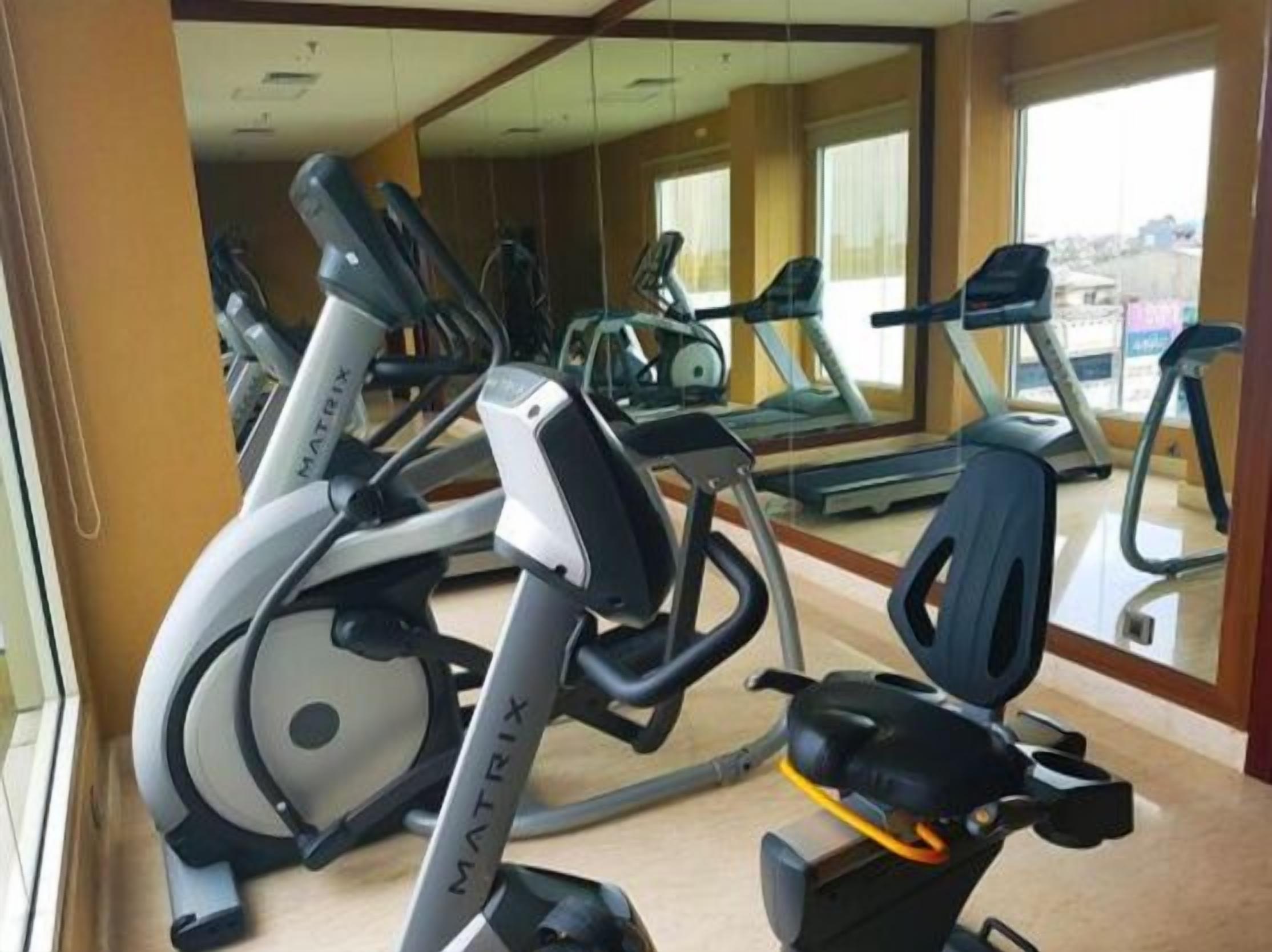 Un gimnasio con varias cintas de correr y bicicletas estáticas, ventanas amplias y un espejo grande.