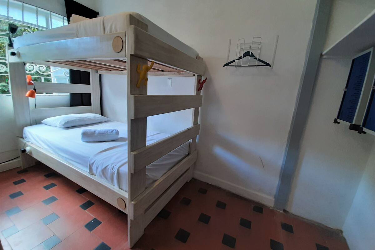 Ein Stockbett mit weißer Matratze und Kissen, ein kleines Regal mit einem roten Gegenstand und ein Wandhaken.