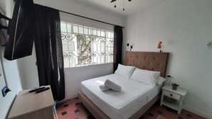 Ein ordentlich eingerichtetes Schlafzimmer mit einem Bett, einem Nachttisch, einem Ventilator an der Decke, einem Fenster mit Metallgitter und einer kleinen Topfpflanze.