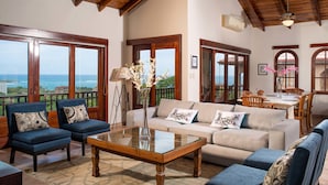 Un vaste espace de vie doté d’un plafond en bois, de grandes fenêtres offrant une vue sur la mer, d’un ensemble de canapés et d’une table à manger avec des chaises.