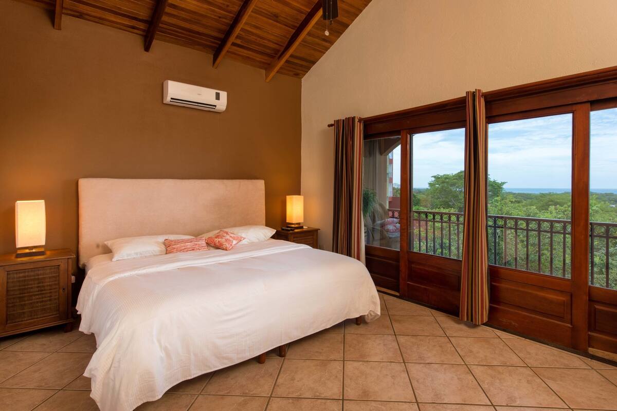 Un dormitorio con una cama grande, cabecera de madera, dos lámparas de noche, una mesita de noche y vistas a zonas verdes a través de una ventana.