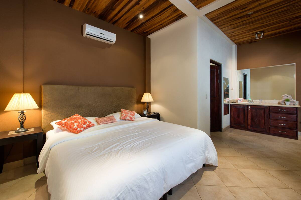 Un dormitorio con una cama grande, dos mesitas de noche con lámparas, techo de madera y un baño con lavamanos y espejo.