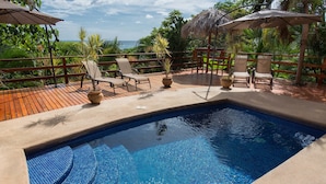 Une piscine entourée d’une terrasse en bois et d’une végétation luxuriante, avec des chaises longues et des parasols à proximité.