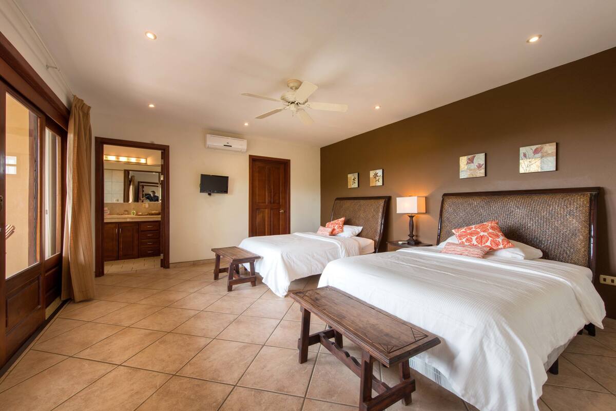 Habitación de hotel con dos camas, ventilador de techo, baño con espejo y vista al exterior.