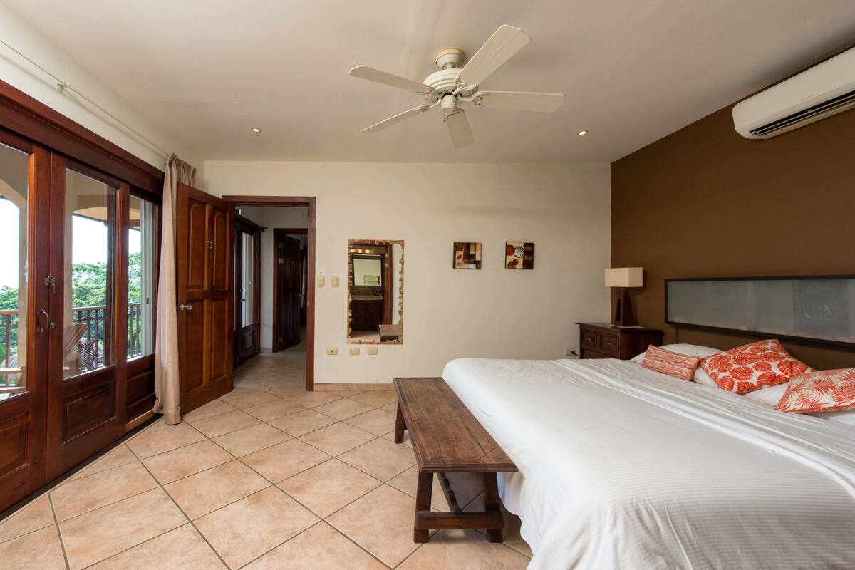 Un dormitorio con cama, ventilador de techo, equipo de aire acondicionado, un banco y vistas a través de una puerta corrediza.