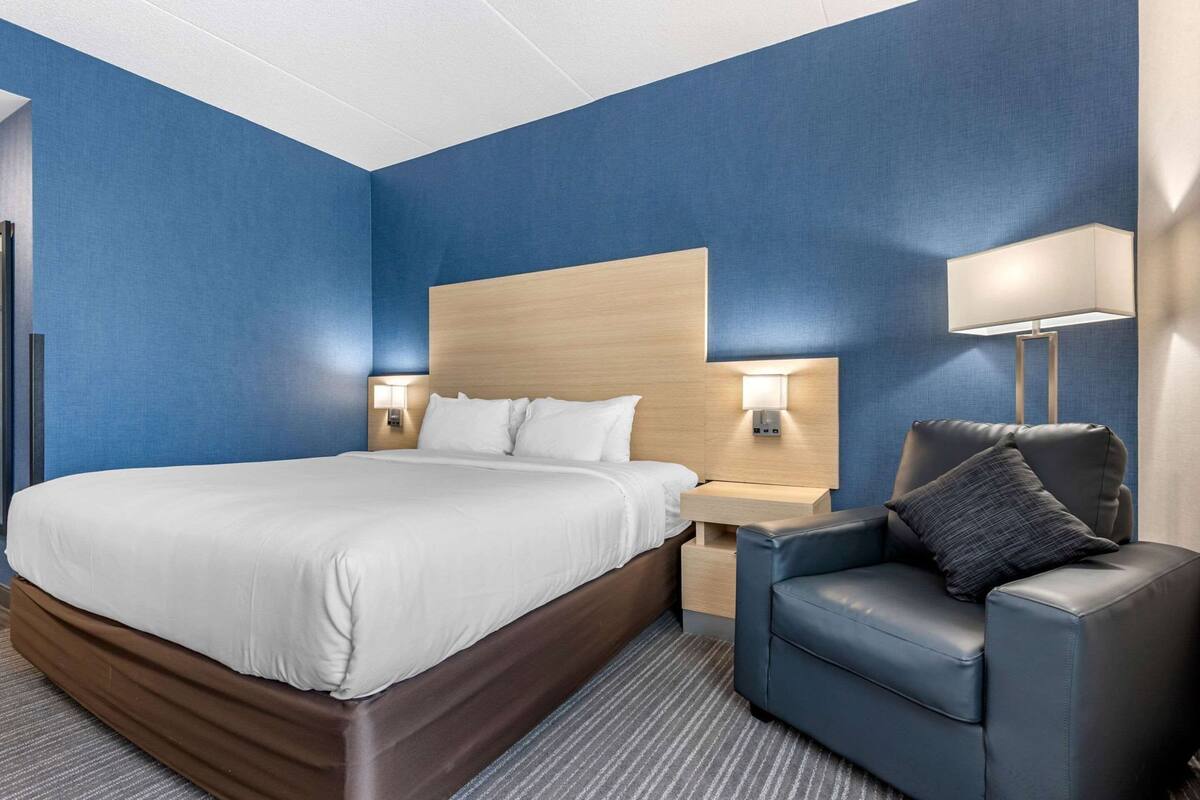 Una habitación de hotel con una cama grande, una mesita de noche con lámpara, una silla y un cabecero.
