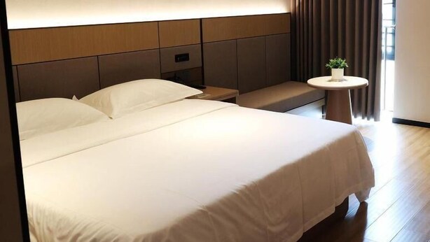 Una habitación de hotel moderna con una cama grande, una mesita de noche, una pequeña mesa redonda con una planta y vistas al exterior a través de cortinas.