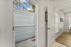 A hallway with white walls, a wooden floor, and a door leading to a covered outdoor area with a view of trees.
